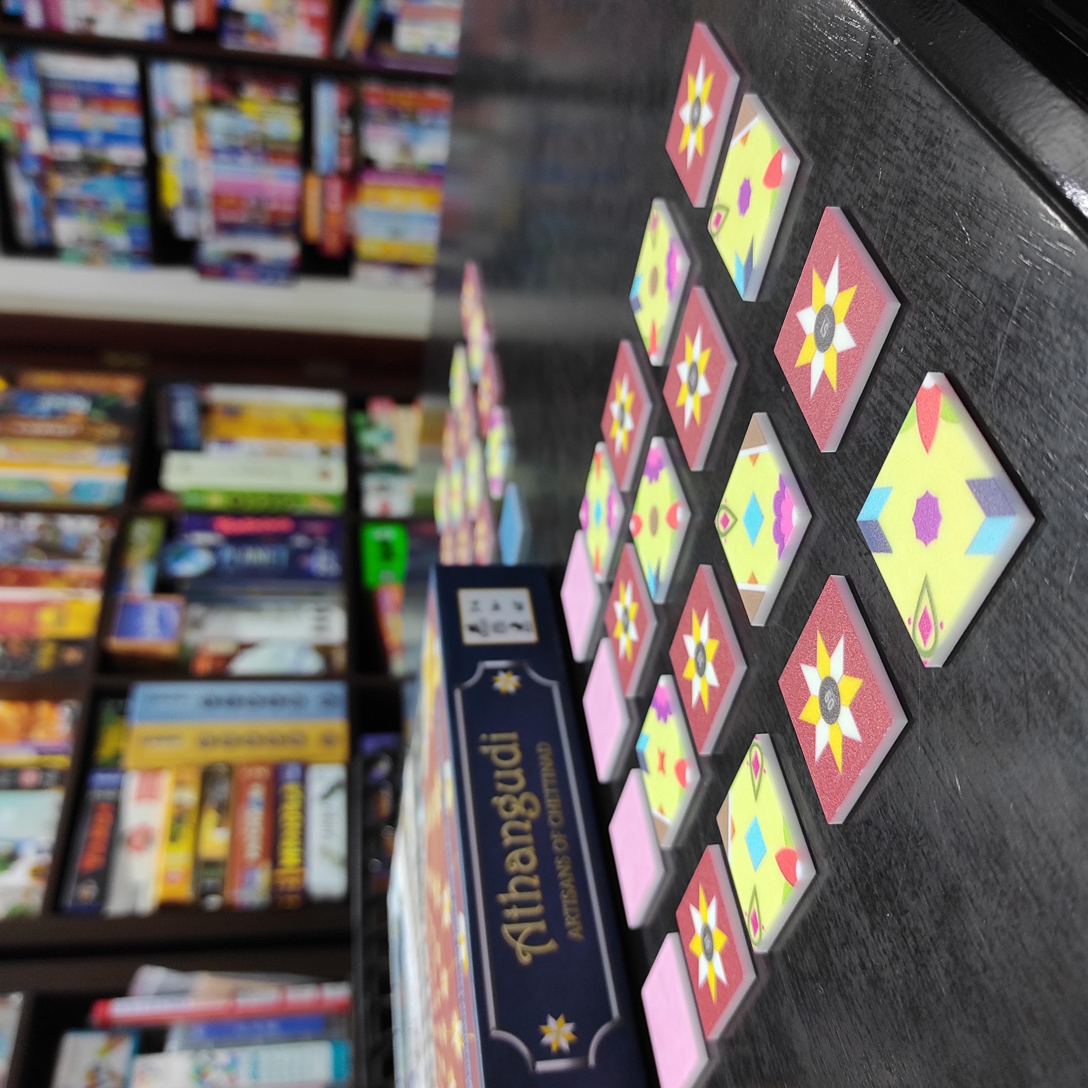 Tiles of the game Athangudi: Artisans of Chettinad, laid out on a table. The game box is visible in the background, as as several shelves filled with boardgames. 