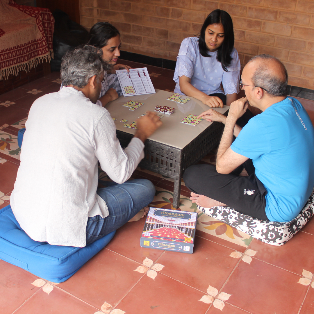 A family of four, seated on the floor, play a game of Athangudi: Artisans of Chettinad that's placed on a low table. The floor is made of Athangudi tiles.