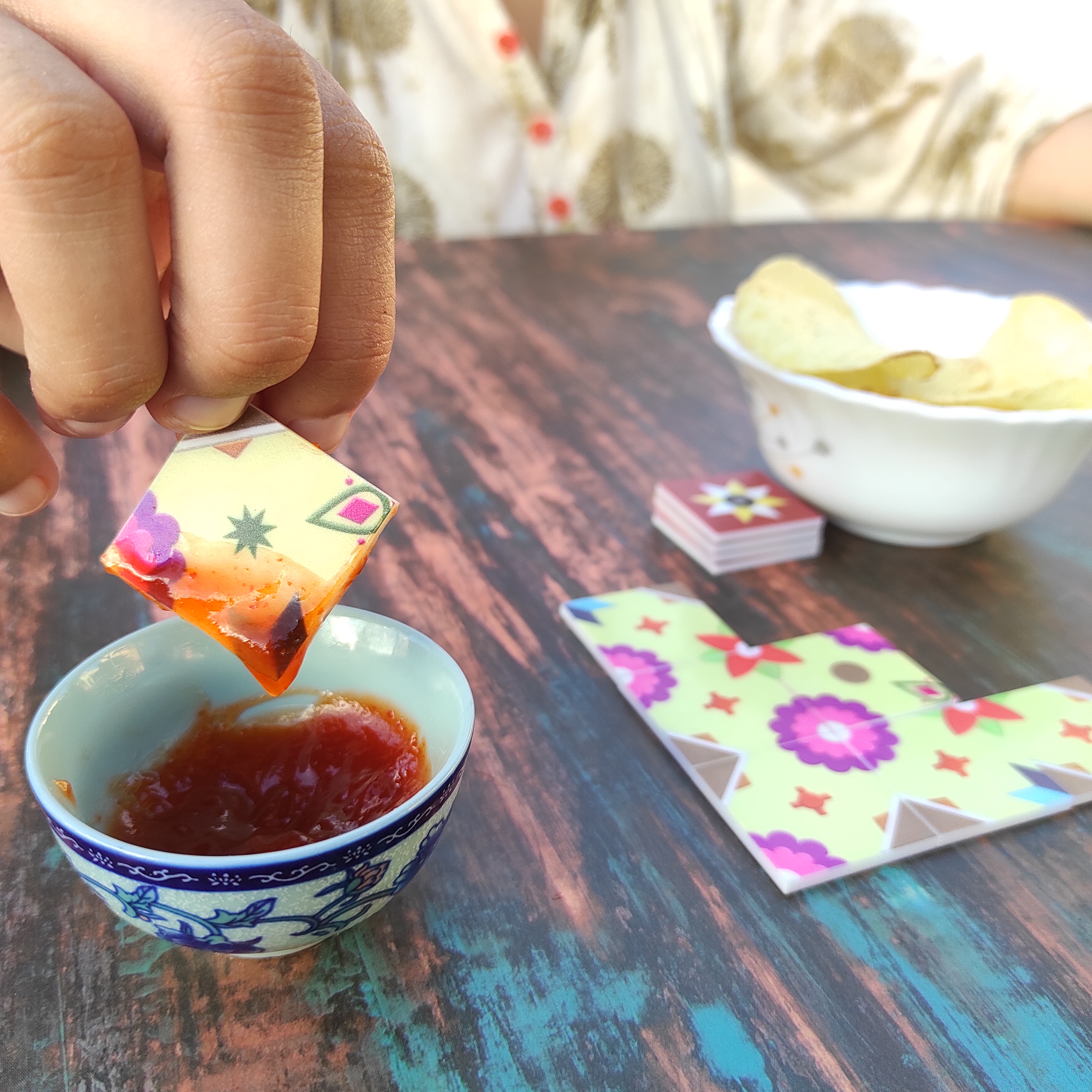 Image of a person dipping a playing tile of the game Athangudi: Artisans of Chettinad into a bowl of Ketchup. The game is going on in the background, and a bowl of potato chips is also seen indicating she might have dipped the tile in the ketchup by mistake instead of the chips. 