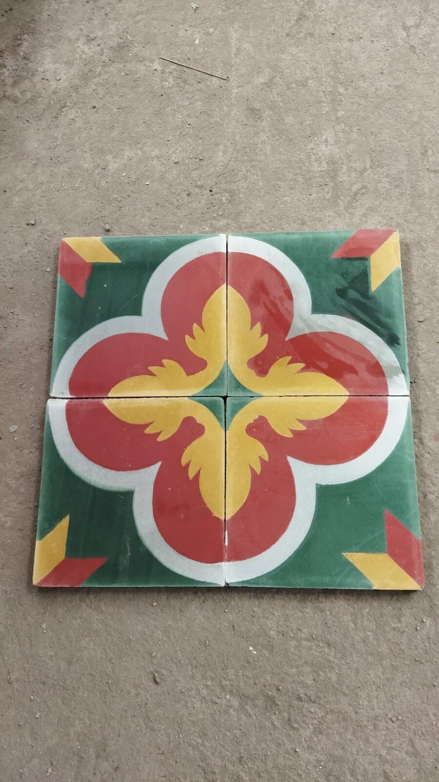 Four Athangudi tiles have been placed on the floor in a 2x2 formation such that they make a big square in which each quarter is one tile. The tiles have a green background, and a flowery motif of red, yellow and white at the center. The corners of the square have a small motif, which looks like a yellow diamond and a red diamond placed next to each other. 