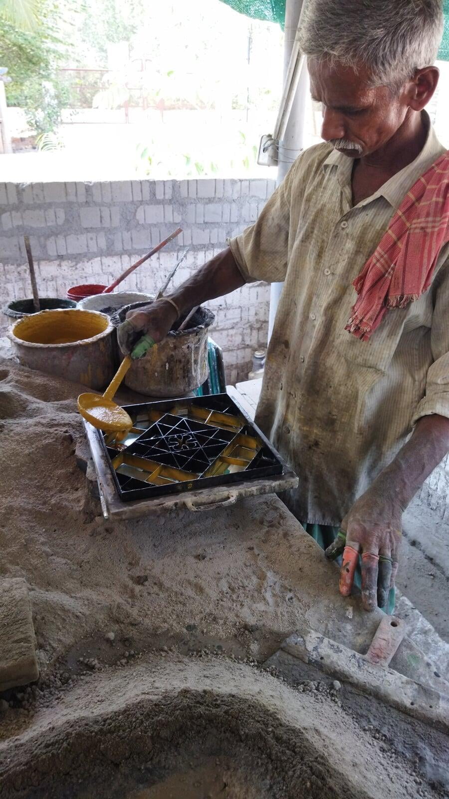An elderly male artisan in the process of making an Athangudi tile. A metal frame containing the pattern of the tile is placed on a platform. The artisan is pouring yellow paint into the frame with a ladle. 