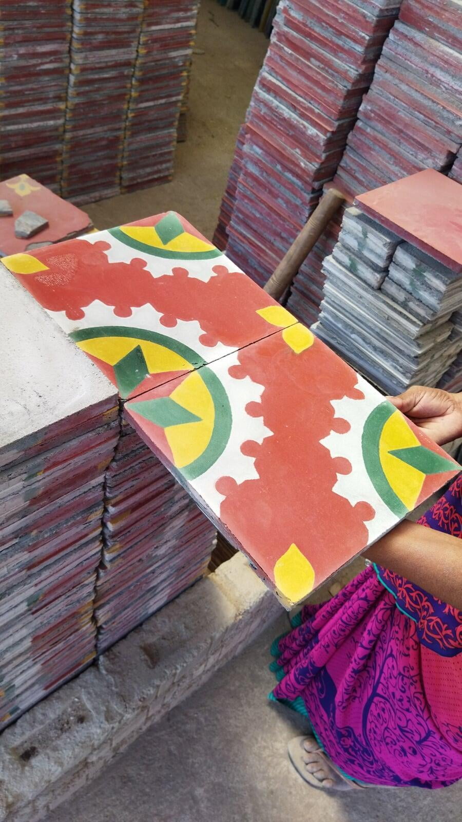 An artisan displays two Athangudi tiles next to each other, so that a partial circular motif is formed by the corners of the two tiles. The tiles are have a red background, and in the corners, a circular pattern surrounding one quarter of an eight pointed star with alternating green and red segments. 