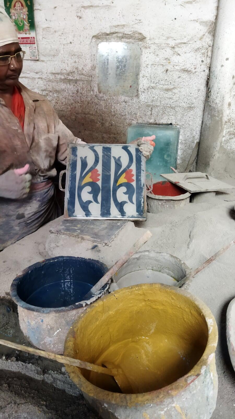 A female artisan displays an Athangudi tile at the end of its creation, but while still in its mold. The tile is divided into halves, and each half has the same pattern - a dark blue vine with a red and yellow flower on a white background. In the foreground of the picture are two buckets of vibrant paint, dark blue and yellow in colour, that have been used to make the tile being displayed. 