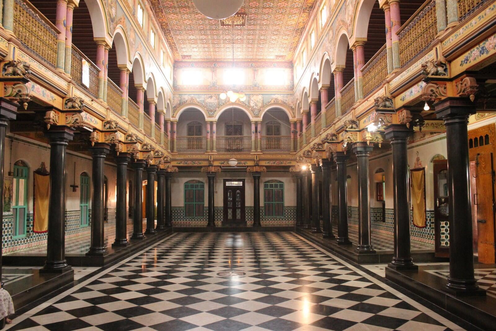 A View of the Grand hall of the "Periya Veedu" (Big House) in Athangudi Village. The photo shows a huge hall with a chequered floor of Italian black & white marble. The floor is flanked by marble pillars which stretch along a corridor. There is an upper floor above the corridor, overlooking into the grand hall. The upper floor has a walkway flanked by colourful pillars and a balustrade. The ceiling of the hall is filled with intricate and colourful geometric designs. Afternoon sunlight streams in through several small windows which are positioned just below the ceiling. 