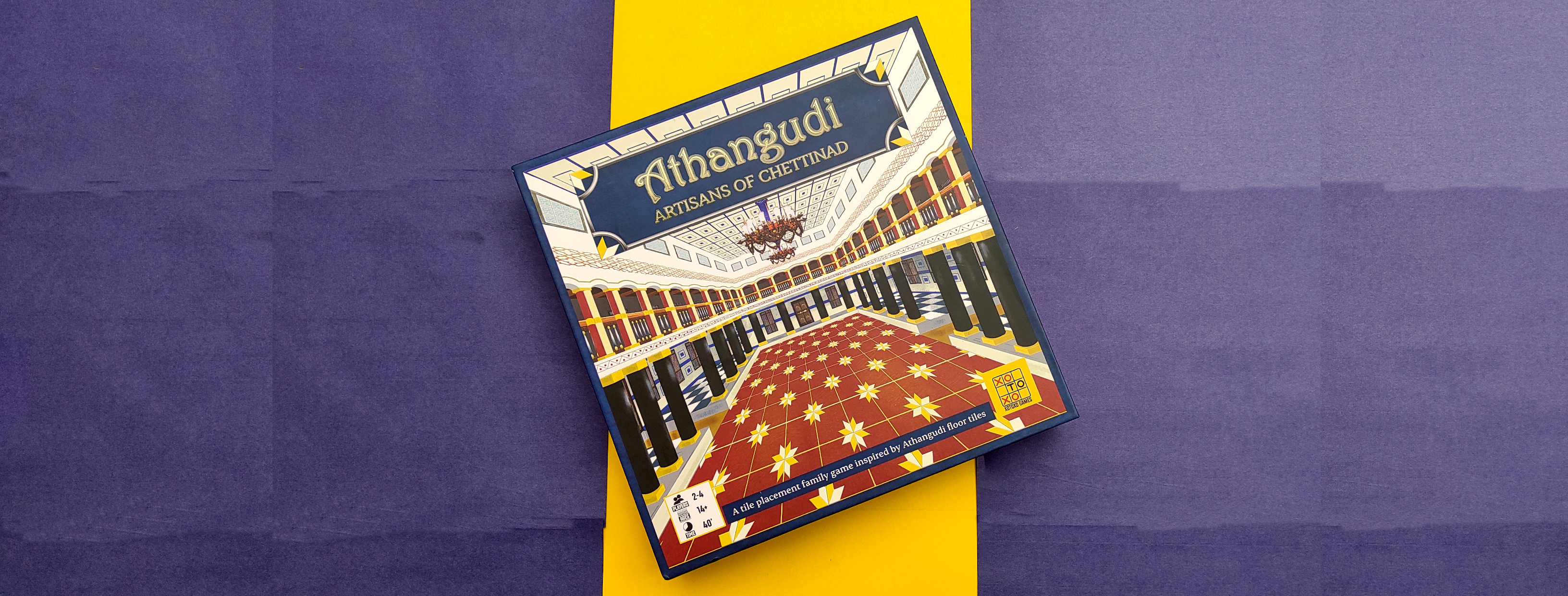 Box of the boardgame Athangudi: Artisans of Chettinad placed at an angle on a background of navy blue and yellow