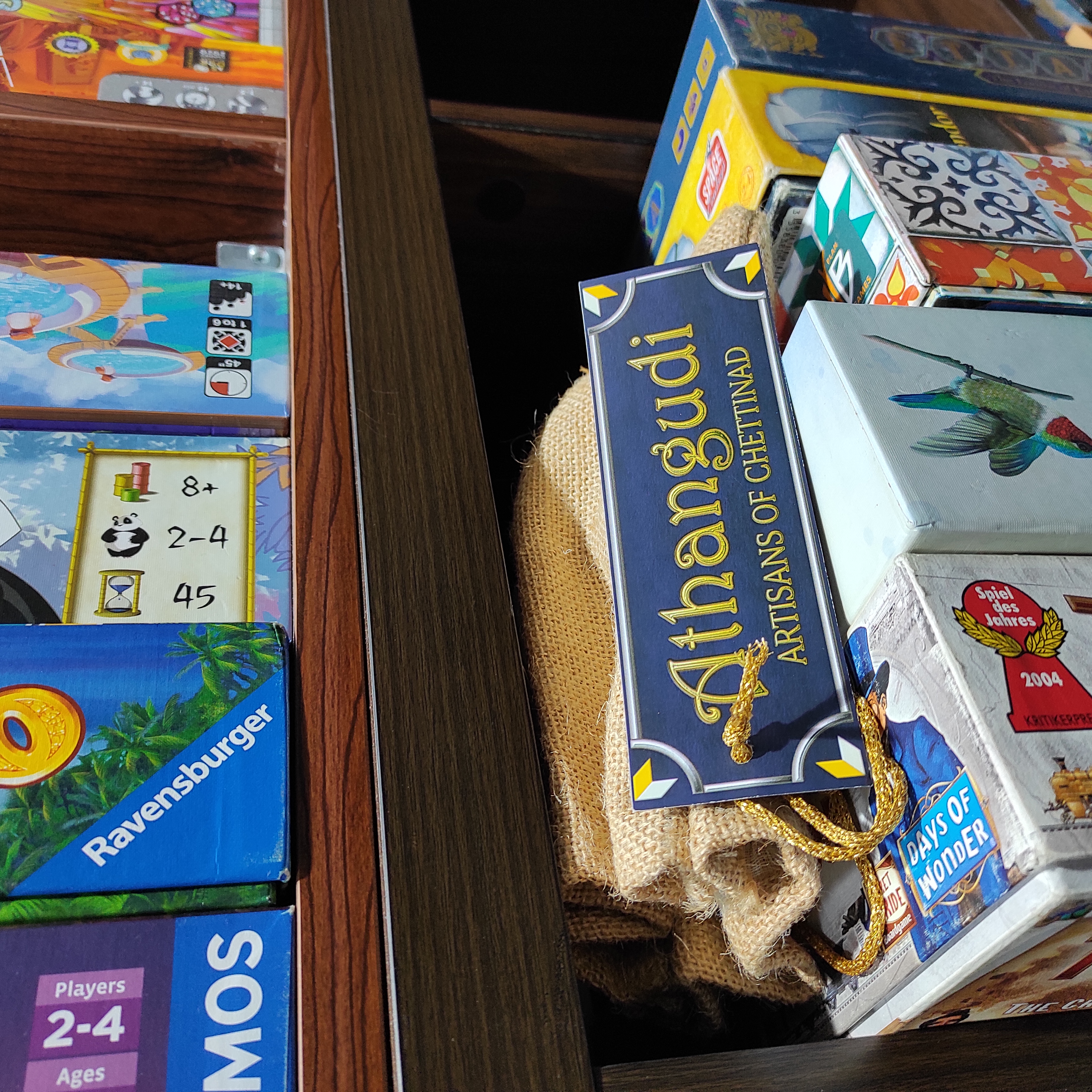 Picture shows a jute pouch with a label displaying "Athangudi: Artisans of Chettinad" tied to a golden drawstring. The pouch is placed in a shelf that's full of several boardgames. The pouch is placed in the little bit of space that's on top of the existing games. 