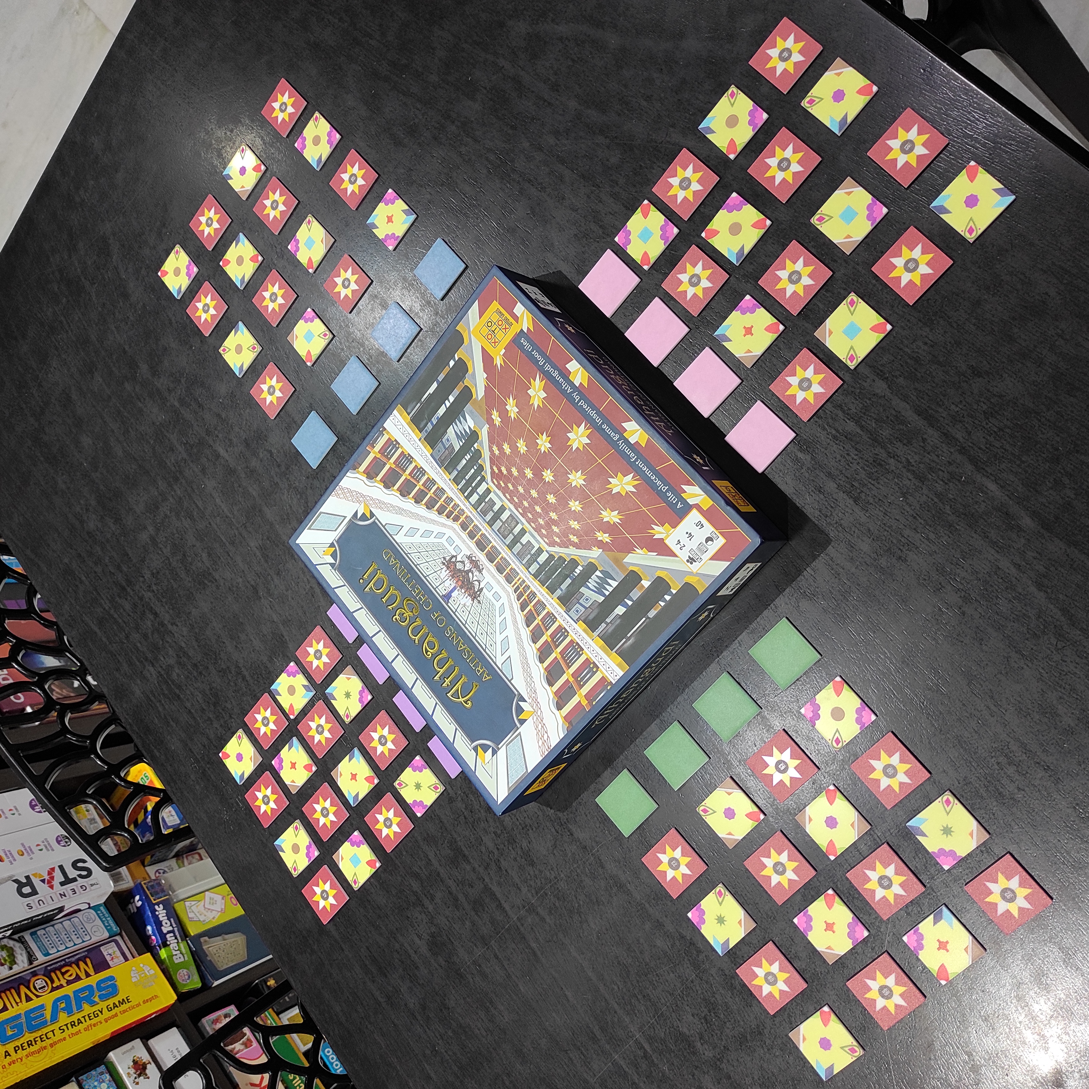 Picture shows the game Athangudi: Artisans of Chettinad laid out on a table in the form of a cross. The Box makes the center of the cross, while tiles from the game are arranged in 4 columns to make up each of the arms of the cross. Some boardgames are visible in the background on a shelf.